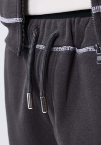 Pantalones de chándal negros con cordón y costuras en overlock blancas, con ojales metálicos plateados en la cintura, combinados con una chaqueta con cremallera a juego.