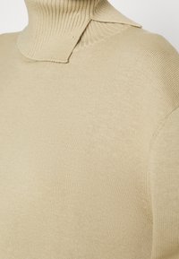 Pull à col roulé beige en tricot avec un détail de col superposé et plié.
