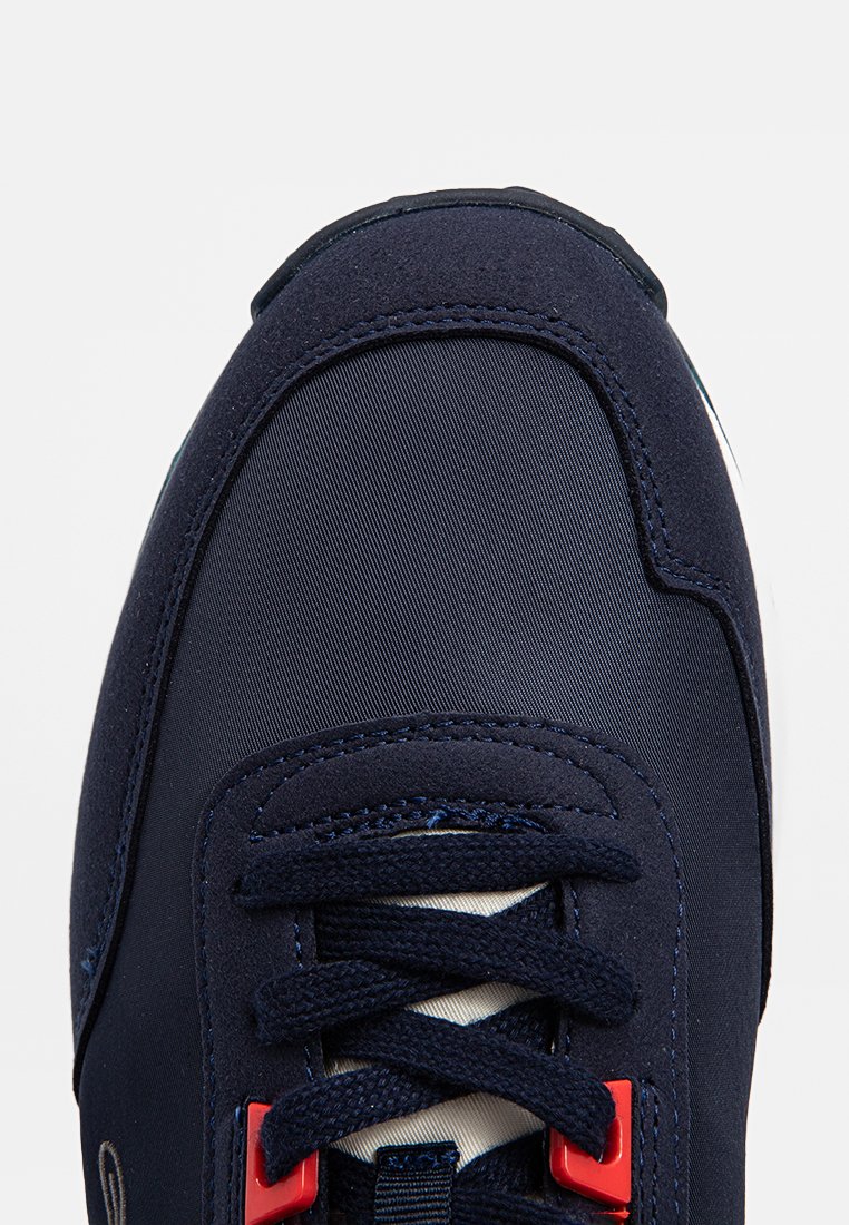 Marinblå sneakers med ovandel i textil, snörningssystem och röda accenter. Har en rundad tå och texturerad yta.