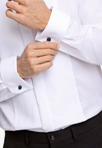 Camicia bianca con tessuto a trama, gemelli neri a contrasto e chiusure a bottoni, indossata con una cintura e pantaloni neri.