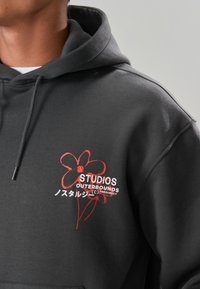 Felpa con cappuccio grigia realizzata in tessuto morbido con cappuccio regolabile con coulisse. Presenta una grafica di un fiore rosso e testo bianco sul lato sinistro del petto.