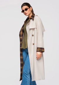 Femme portant un trench-coat beige avec doublure à carreaux, un haut vert, un jean bleu et de petites lunettes de soleil foncées, debout devant un fond uni.