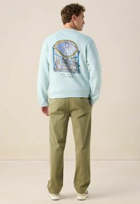 Sweatshirt bleu clair avec un imprimé graphique d'une galerie d'art et du texte dans le dos. Assorti avec un pantalon vert olive et des baskets blanches.