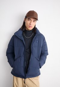 Marineblå puffjakke med glidelås, sidelommer og hette. Båret over en svart genser med høy hals, kombinert med en brun caps.