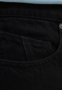 Zwarte denim jeans met een klein voorvak, voorzien van donkere stiksels en een zilveren knoopdetail. De stof lijkt soepel met een lichte textuur.