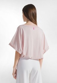 Camiseta rosa clara de gran tamaño en tela suave, con mangas remangadas y una pequeña etiqueta rosa en la parte trasera. Se lleva con pantalones blancos de pierna ancha.