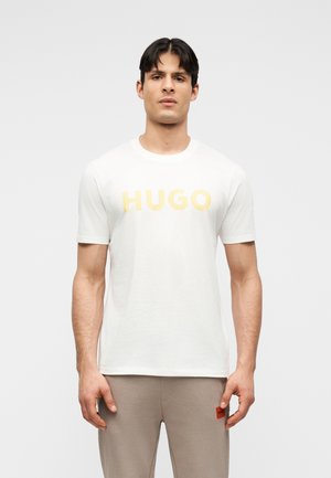 Ein Mann steht nach vorne gerichtet und trägt ein weißes T-Shirt mit der Aufschrift "HUGO" in Gelb sowie beige Hosen mit einem kleinen orangefarbenen Logo-Patch.