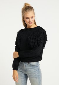 Pulover negru tricotat, cu detalii cu franjuri pe umeri, decolteu rotund și manșete cu model texturat. Purtați cu blugi albaștri deschisi.