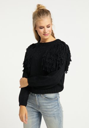 Maglione nero a maglia con dettagli di frange sulle spalle, scollatura rotonda e polsini a costine. Indossato con jeans azzurri chiari.