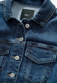 Veste en jean bleu foncé avec des boutons métalliques étiquetés "NEXT JEANS SINCE 1982" et une étiquette de col noire avec "NEXT" en lettres blanches.