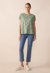 Grön bomullströja med korta ärmar och rundad halsringning, prydd med ett spritt bladmönster. I kombination med blå rak-linjes jeans och vita sneakers.