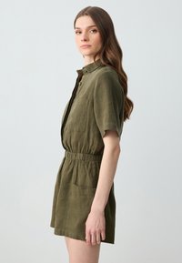 Olivgrüner Leinen-Jumpsuit mit kurzen Ärmeln, Knopfleiste, elastischem Bund und einer Brusttasche. Leichter, strukturierter Stoff.