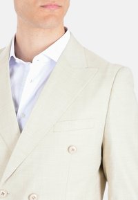 Blazer beige doppiopetto realizzato in tessuto testurizzato, con rever a punta, taschino sul petto e bottoni di colore chiaro. Camicia bianca sotto.