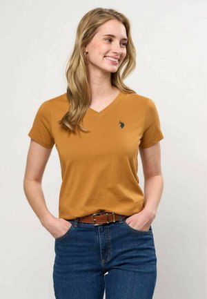 Jeune femme aux longs cheveux blonds, portant un t-shirt jaune moutarde à col en V et un jean bleu avec une ceinture marron, souriante et regardant sur le côté.