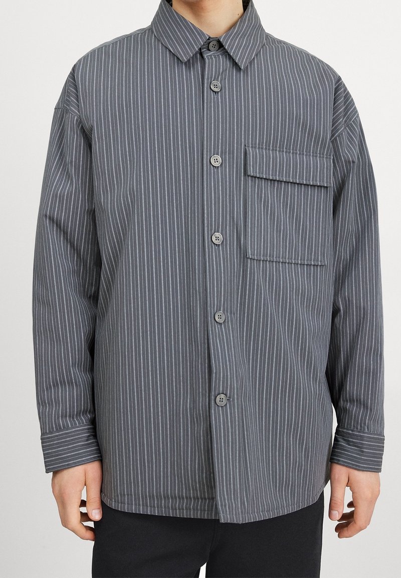 Homme portant une chemise grise à manches longues boutonnée avec de fines rayures blanches verticales et une grande poche poitrine avec rabat.