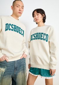 Dvaja ľudí majú na sebe krémové mikiny s nápisom "DCSHOECO" v teal farbe. Jeden z nich ich kombinuje s poškodenými modrými džínsami, zatiaľ čo druhý má teal šortky s čipkovým lemom.