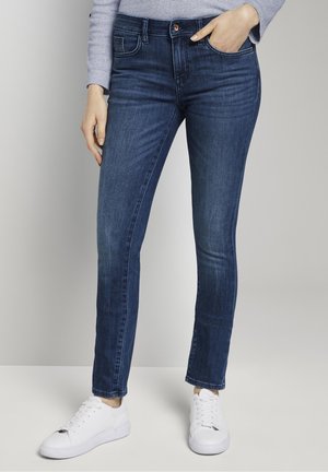Femme portant un jean skinny bleu marine, un haut à manches longues gris clair et des baskets blanches, debout avec une main dans la poche.