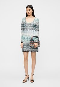 Φόρεμα με μακριά μανίκια που διαθέτει πολύχρωμο κυματιστό σχέδιο σε ανοιχτό μπλε και μαύρο, συνδυασμένο με μικρή τσάντα χειρός με μοτίβο και μαύρα πέδιλα με λουράκια.