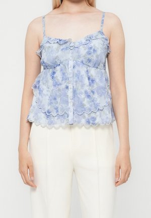 Femme portant un top bleu fleuri sans manches avec volants et boutons, associé à un pantalon blanc taille haute sur fond uni.