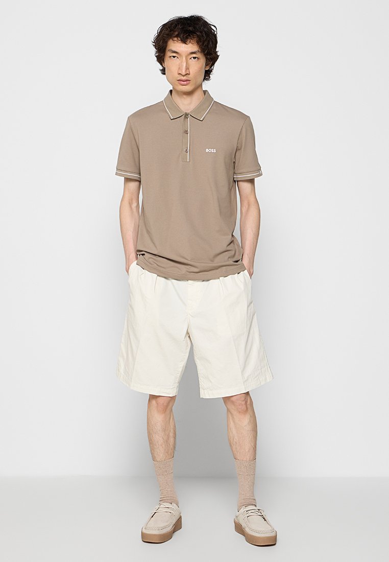 Beige Polo-Shirt mit kurzen Ärmeln, Kragen und gesticktem Logo, kombiniert mit cremefarbenen Shorts und hellen Schuhen.