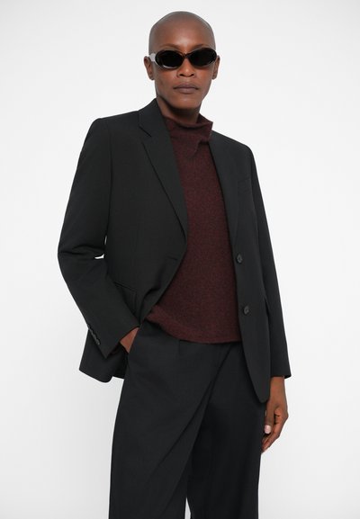 Must tikkivad bleiser musta turtlenecki peal, koos mustade pükstega ja ümmarguste mustade prillidega, luues sujuva monokroomse ilme.
