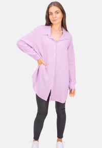 Hellpurpurnes Hemdkleid aus strukturiertem Stoff mit Kragen, langen Ärmeln und seitlichen Taschen. Kombiniert mit schwarzen Leggings und weißen Sneakers.