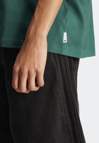 T-shirt verde in cotone con orlo cucito e una piccola etichetta bianca del marchio, abbinata a pantaloncini neri testurizzati con strisce laterali.
