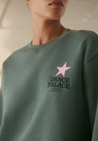 Sudadera verde hecha de tela suave, con un bordado de "DANCE PALACE" y un gráfico de estrella rosa en el lado izquierdo del pecho.