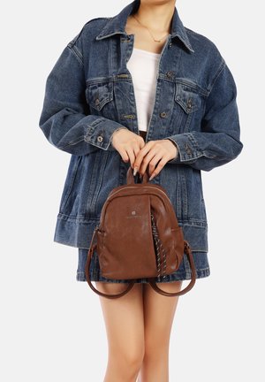 Zaino in pelle marrone dalla forma arrotondata, con doppi manici superiori e un accento a righe bianche e nere. Indossato con una giacca di jeans.