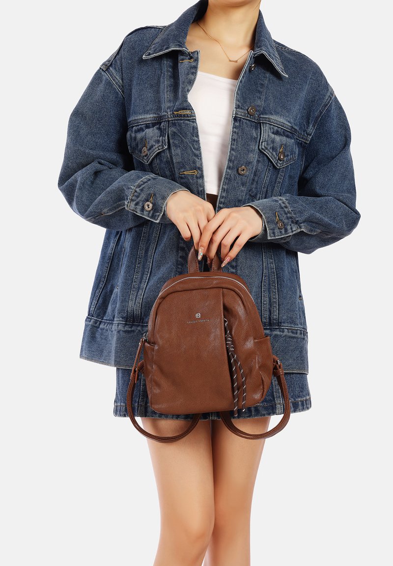 Zaino in pelle marrone dalla forma arrotondata, con doppi manici superiori e un accento a righe bianche e nere. Indossato con una giacca di jeans.