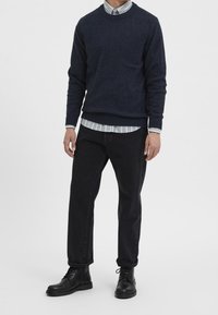 Marinblå stickad tröja över en vit och ljusblå randig skjorta, kombinerad med svarta jeans och svarta anklarstövlar för en avslappnad look.