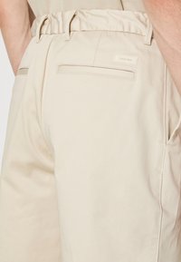Pantalon beige taille haute avec poches passepoilées à l’arrière et petite étiquette avec logo Calvin Klein sur la poche droite, porté par une personne.
