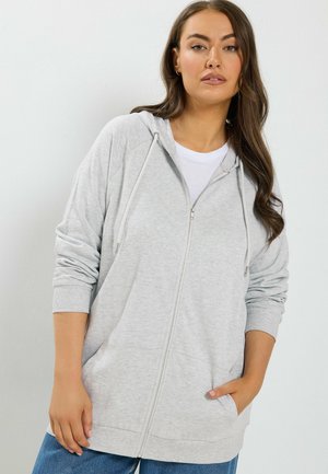 Donna con lunghi capelli castani che indossa una felpa con cappuccio grigio chiaro con zip sopra una maglietta bianca, con una mano in tasca, su uno sfondo bianco uniforme.