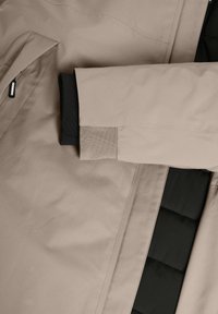 Chaqueta beige con material exterior suave, forro interior negro en contraste y puños de tejido acanalado; incluye un detalle de bolsillo con cremallera.