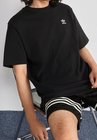 Sort bomuld t-shirt med korte ærmer, der har et lille hvidt Adidas-logo på venstre bryst. Sammen med mønstrede sorte shorts og en stol.