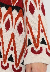 Stickad tröjsleeve med ett geometriskt mönster i varma toner av röd, orange och svart på en krämfärgad bakgrund. Texturerad tygdetalj synlig.