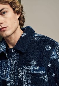 Marineblauwe fleecejack met een gestructureerde afwerking, voorzien van een kraag en een patchzak, versierd met een wit bloemmotief.