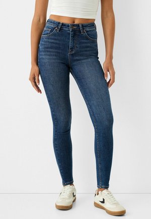 Femme portant un jean skinny bleu foncé taille haute, associé à des baskets blanches et un top blanc court, debout devant un fond uni.