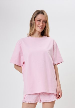 Femme aux cheveux blonds portant un t-shirt rose ample et un short rose à volants assorti, debout devant un fond clair uni.