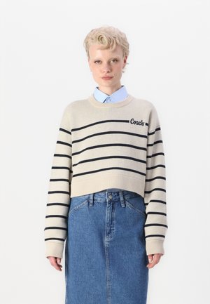 Beige avkortet genser med marineblå striper og "Coach"-logo, kombinert med en blå skjorte med krage og høy midje denimskjørt.