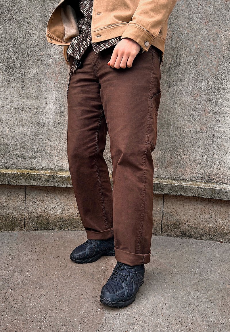 Braune Baumwollhosen mit geradem Bein, kombiniert mit schwarzen Sportschuhen. Der Look wird durch ein gemustertes, strukturiertes Hemd unter einer hellbraunen Jacke ergänzt.