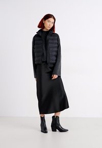 Zwarte puffer vest met horizontale stiksels, gelaagd over een donkere gebreide top en een lange zwarte satijnen rok, gecombineerd met zwarte enkellaarsjes.