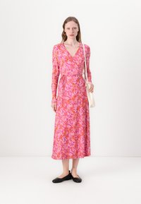 Rochie lungă cu mâneci în roz, cu modele florale în portocaliu și roz deschis. Dispune de o talie legată și o siluetă fluidă, purtată împreună cu pantofi negri.