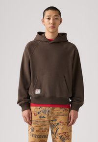 Felpa marrone con tasca frontale, orlo a coste e cappuccio. Indossata sopra una maglietta rossa, abbinata a pantaloni beige a fantasia.