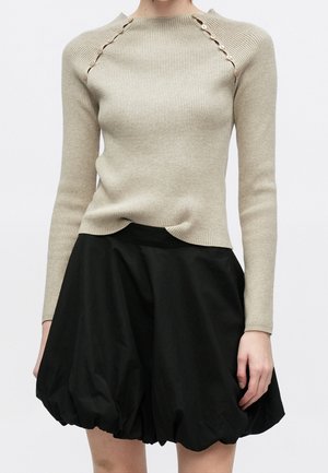 Femme portant un pull en maille côtelée beige avec des détails à boutons sur les épaules et une jupe bulle noire volumineuse.