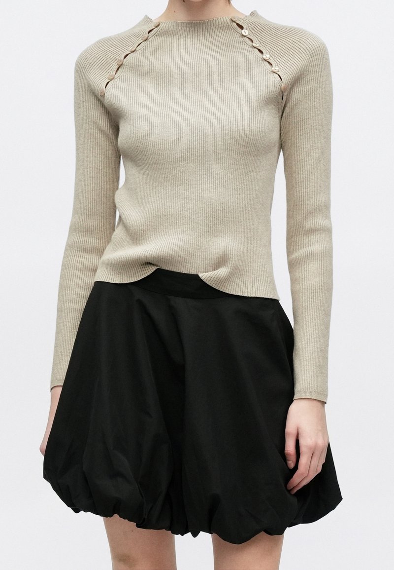 Femme portant un pull en maille côtelée beige avec des détails à boutons sur les épaules et une jupe bulle noire volumineuse.