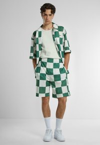 Camicia a maniche corte a quadri verdi e bianchi con pantaloni corti abbinati, dotati di una vita elastica e tasche laterali. Sneakers bianche e calzini.