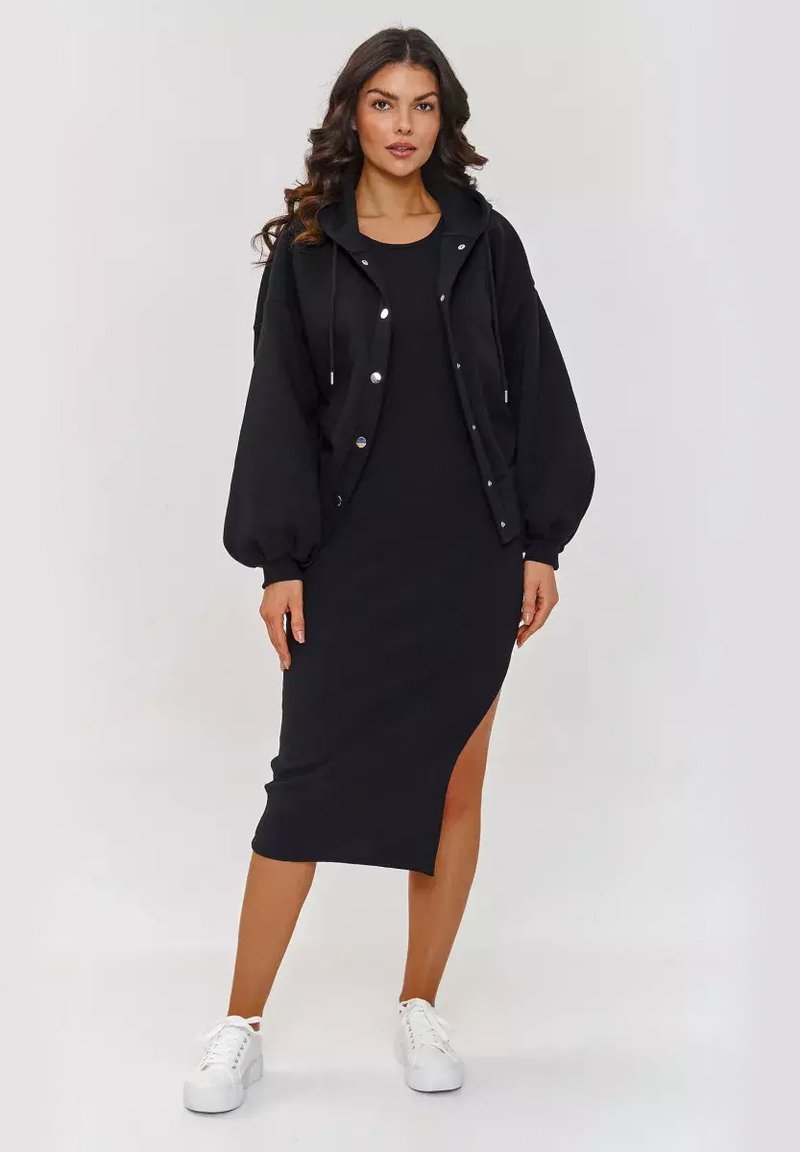 Czarna bomberka z kapturem i zatrzaskami w połączeniu z dopasowaną czarną sukienką do kolan z bocznym rozcięciem, uzupełnioną o białe sneakersy.