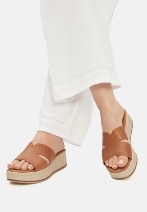 Barna bőr talpbetétes szandálok kivágott mintával és bézs talppal, lekerekített orral és sima textúrával. Fehér nadrággal viselve.