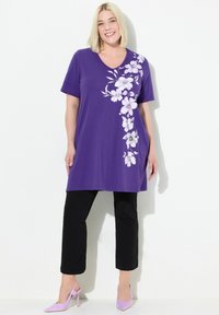 Lila tunika med V-hals och korta ärmar, med vita blommönster på ena sidan; kombinerad med svarta cropped-byxor och lavendelklackar.